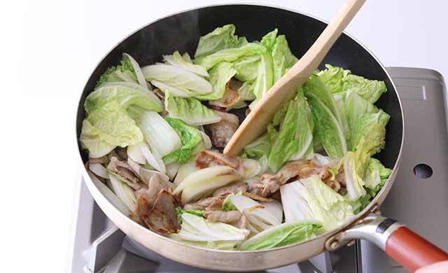 生姜・豚肉・白菜の芯を炒めているフライパンに、白菜の葉と長ねぎを加えて炒めている写真